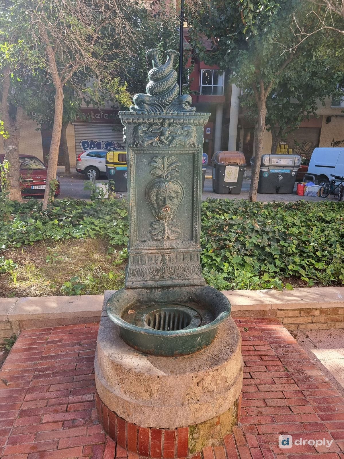 Close-up of a green cast-iron drinking fountain in Benimaclet, Valencia, showing the bearded face with the spout in its mouth, the palmette above it and two intertwined dolphins on top