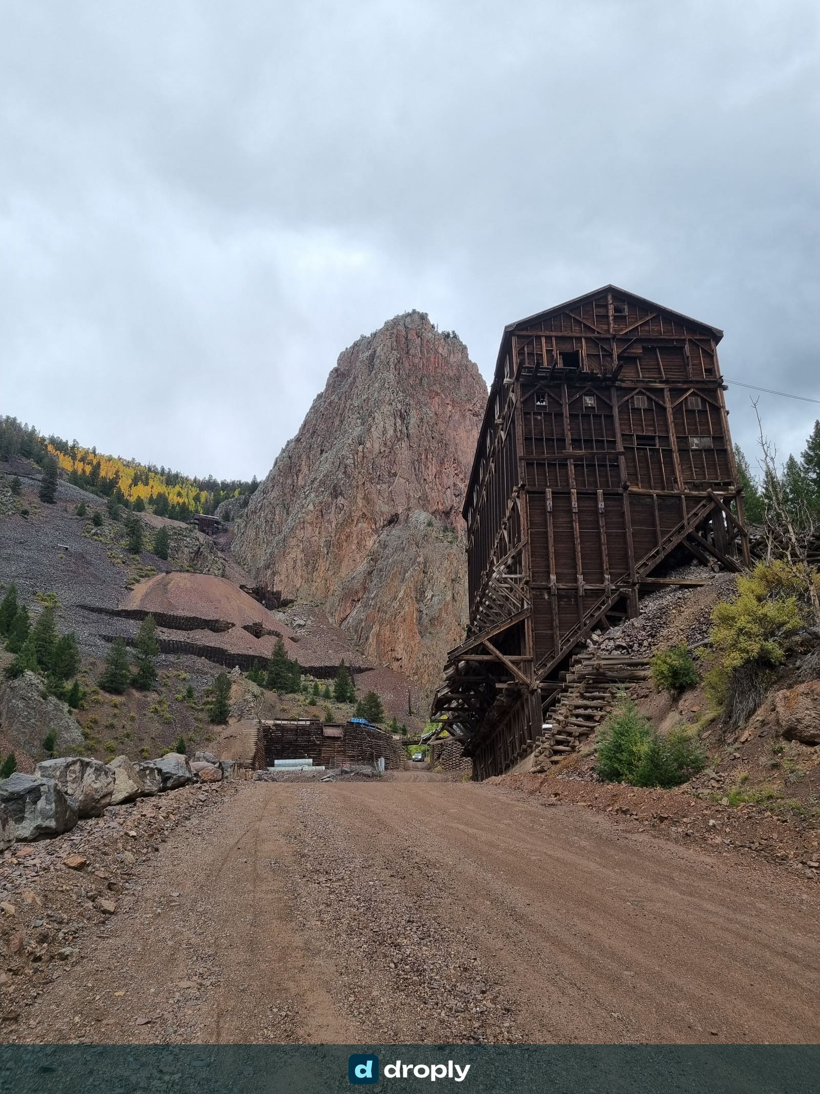 A large wooden mine building on a mountain road in Colorado with a dramatic rock spire behind