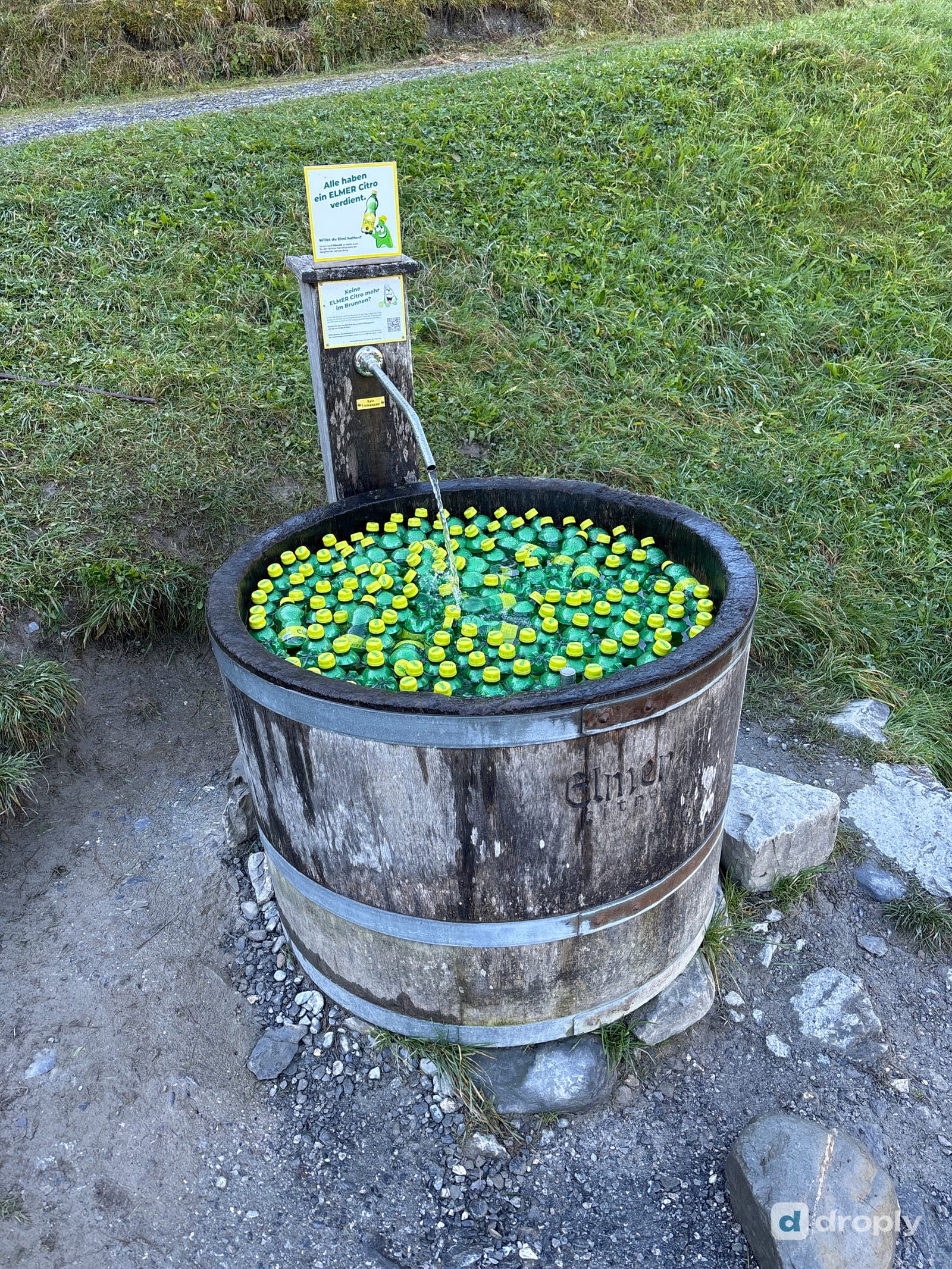 A wooden barrel fountain filled with green Elmer Citro bottles cooling in the running water, above Elm in Glarus