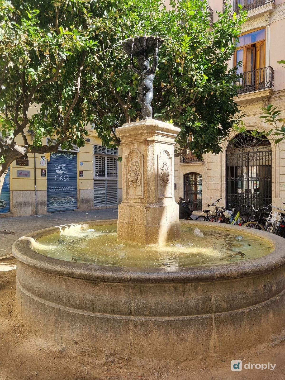 The Font del Negret in Valencia, a child figure holding up a shell on a stone pedestal with sculpted faces on its sides, above a round stone basin