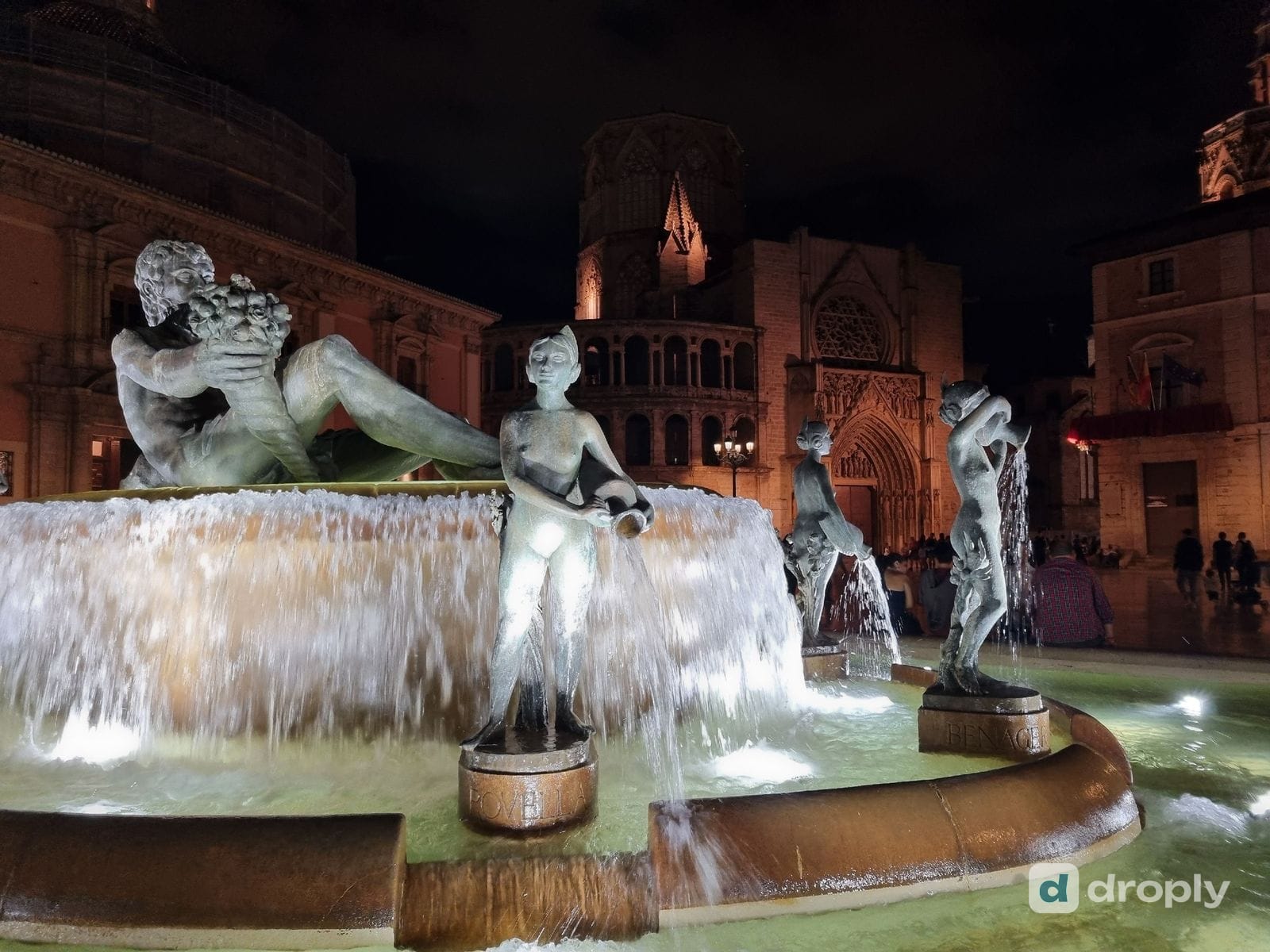 The Turia fountain in Plaça de la Mare de Déu at night, with the bronze river god holding a horn of plenty and the figures Rovella and Benager pouring water from jugs, the Gothic Apostles' door of Valencia Cathedral behind