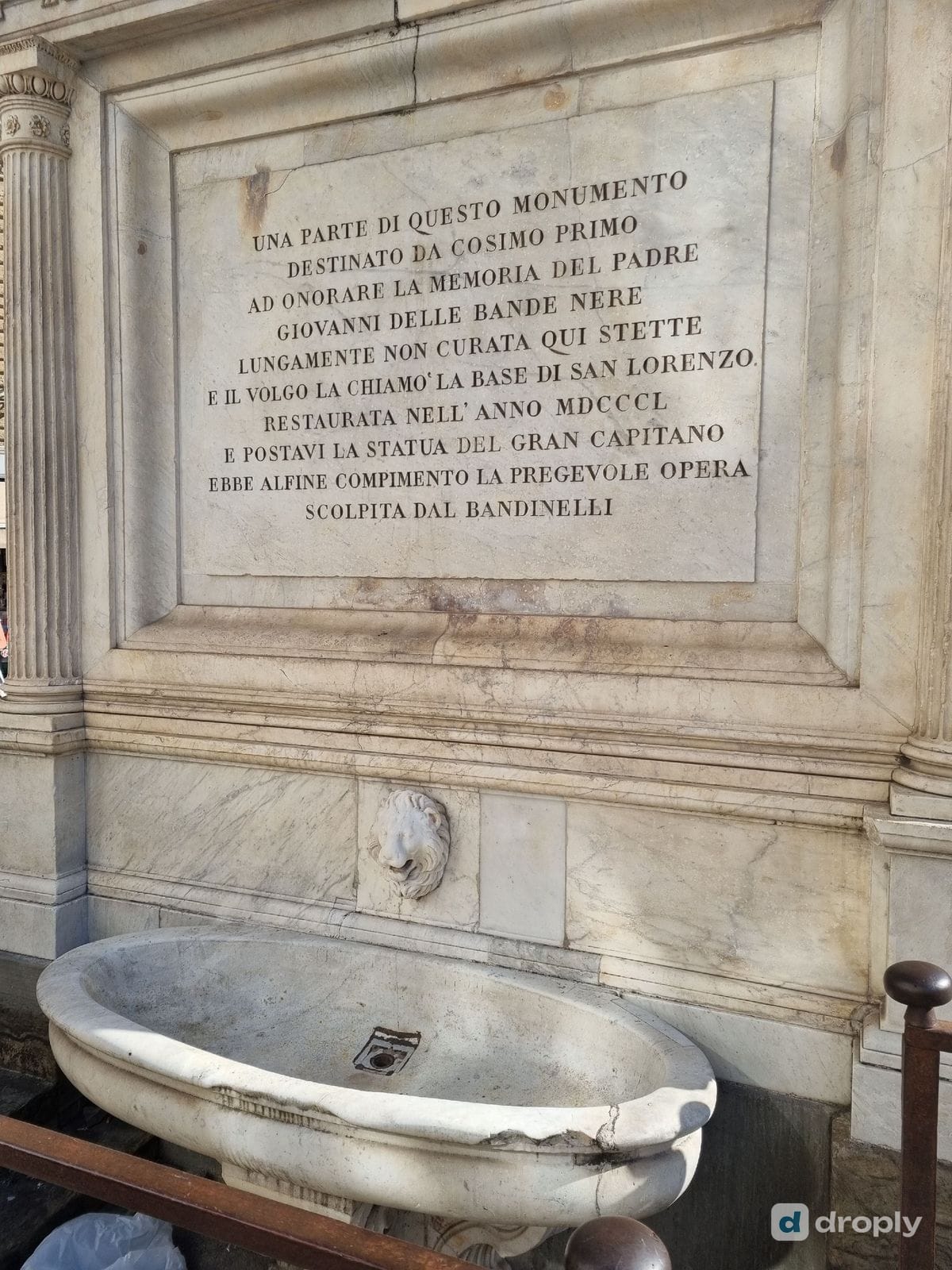 A historic stone monument fountain built into a wall near San Lorenzo in Florence, with an inscription dedicated to Cosimo I de' Medici