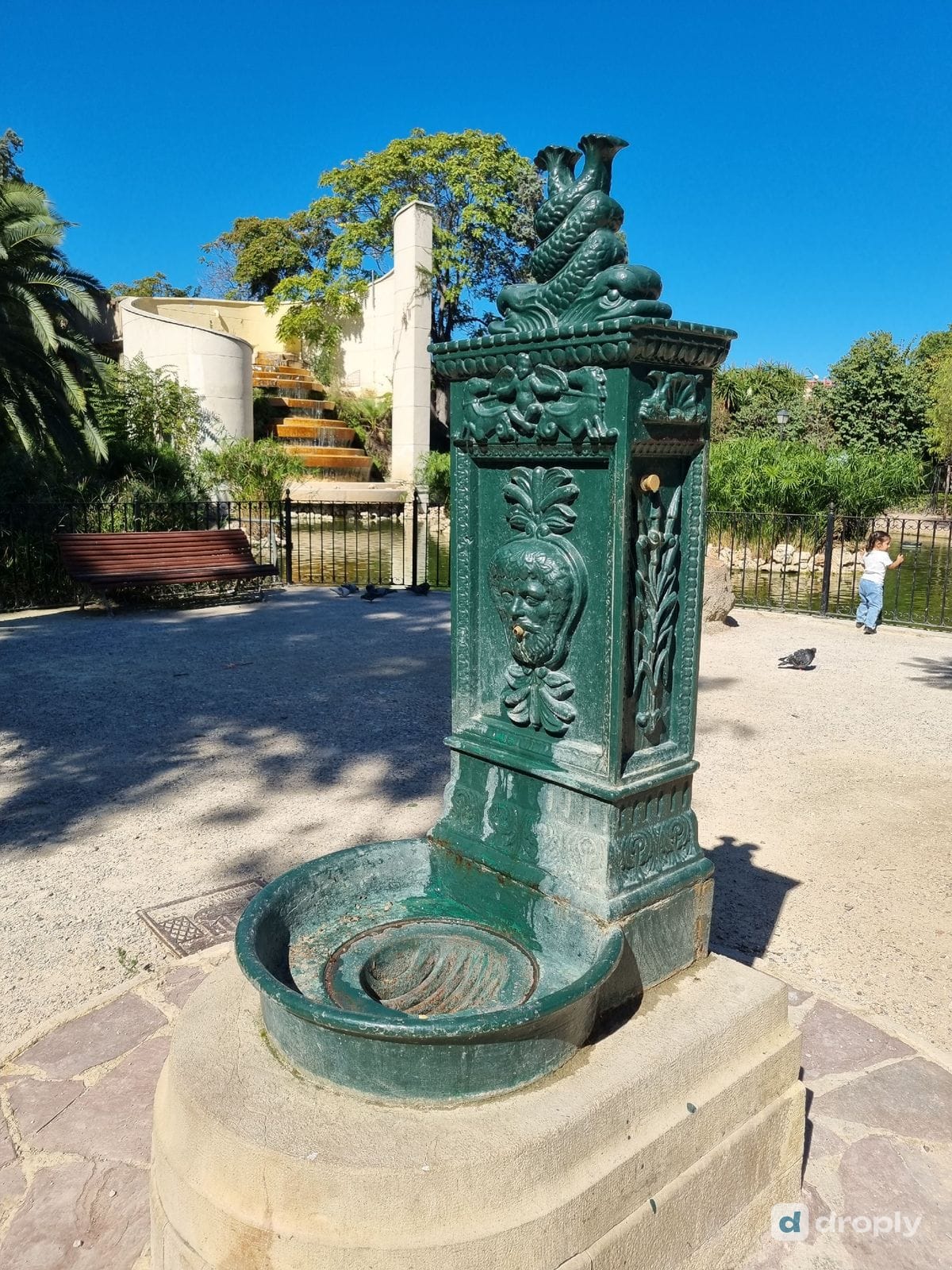 A green cast-iron drinking fountain with a bearded face spout and two dolphins on top, in the Turia Garden in Valencia