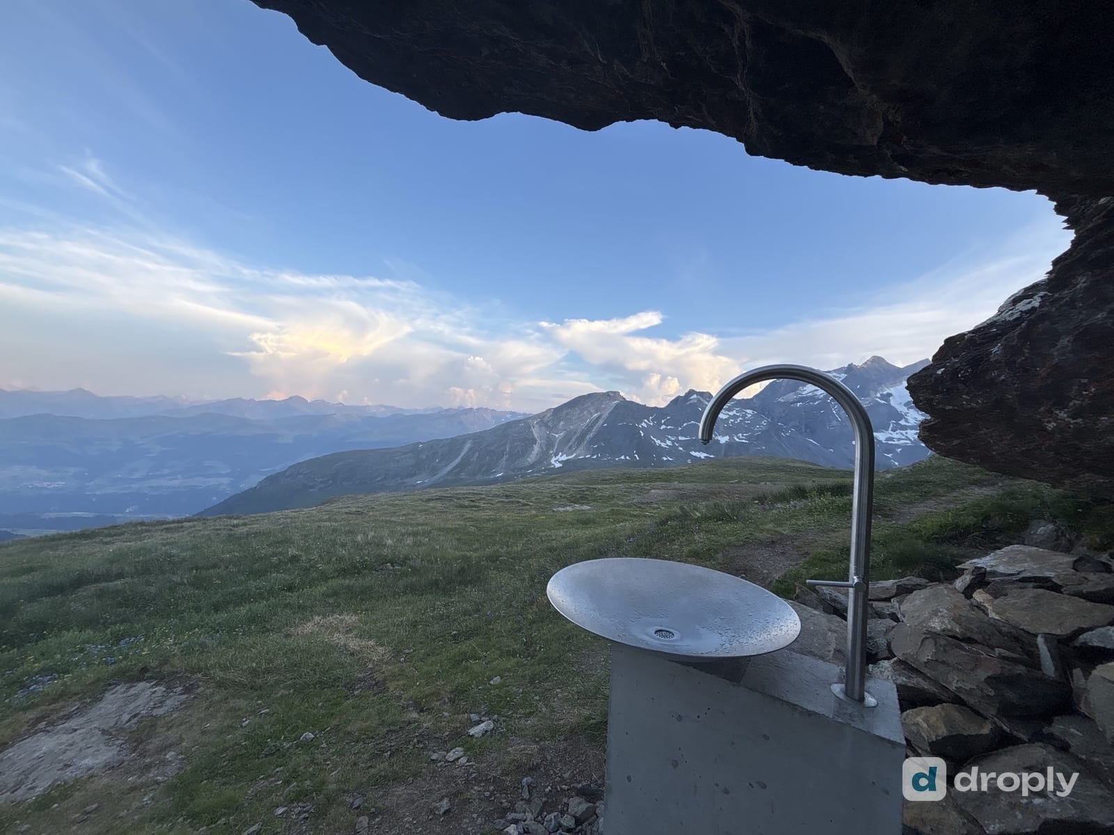 A stainless steel drinking fountain under a rock overhang above Breil/Brigels in Graubünden, Switzerland, with snow-streaked peaks behind it