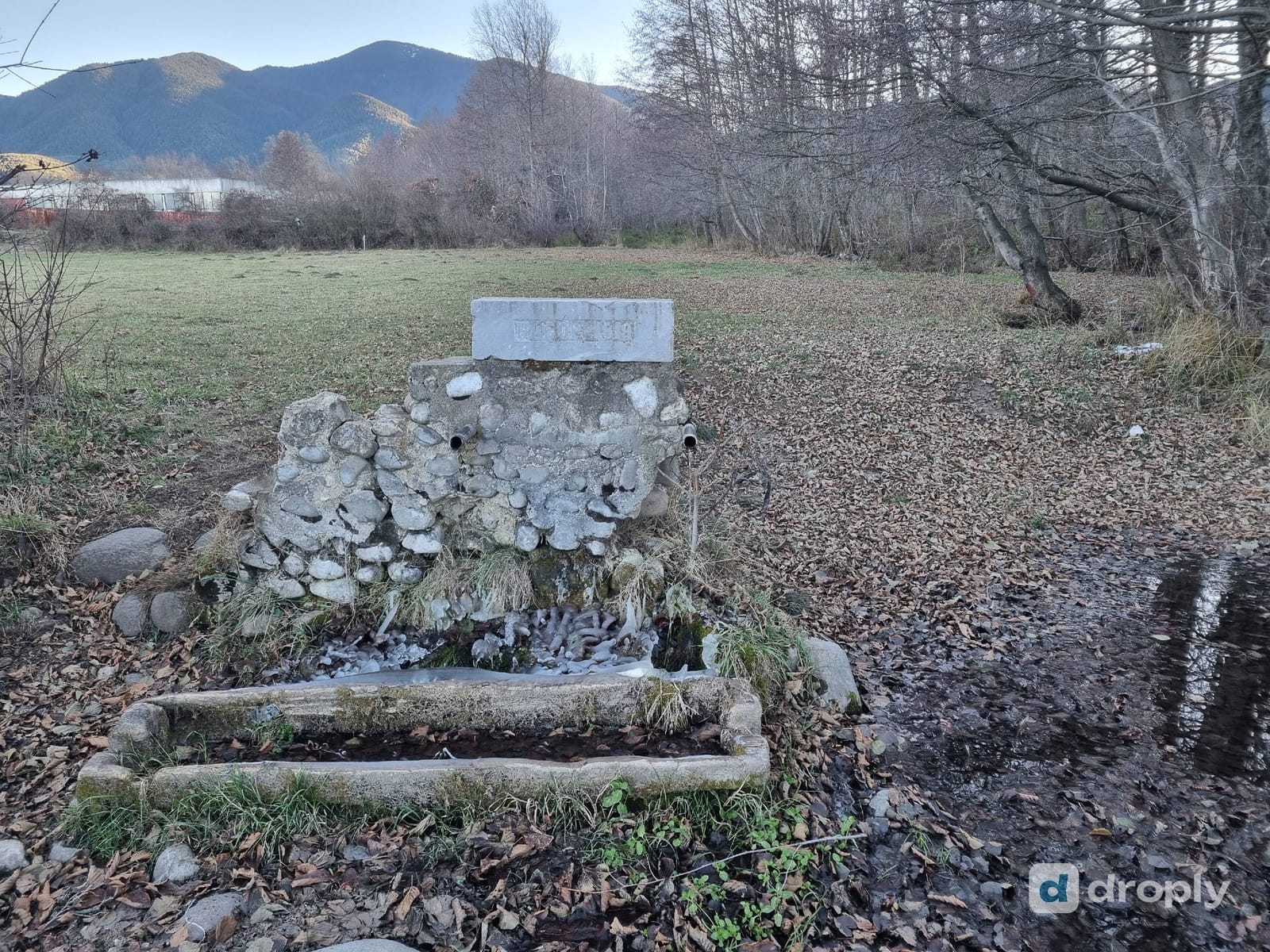 Stone-built spring fountain with a trough near Bansko in the Pirin mountains, partly frozen over, with forested mountains behind