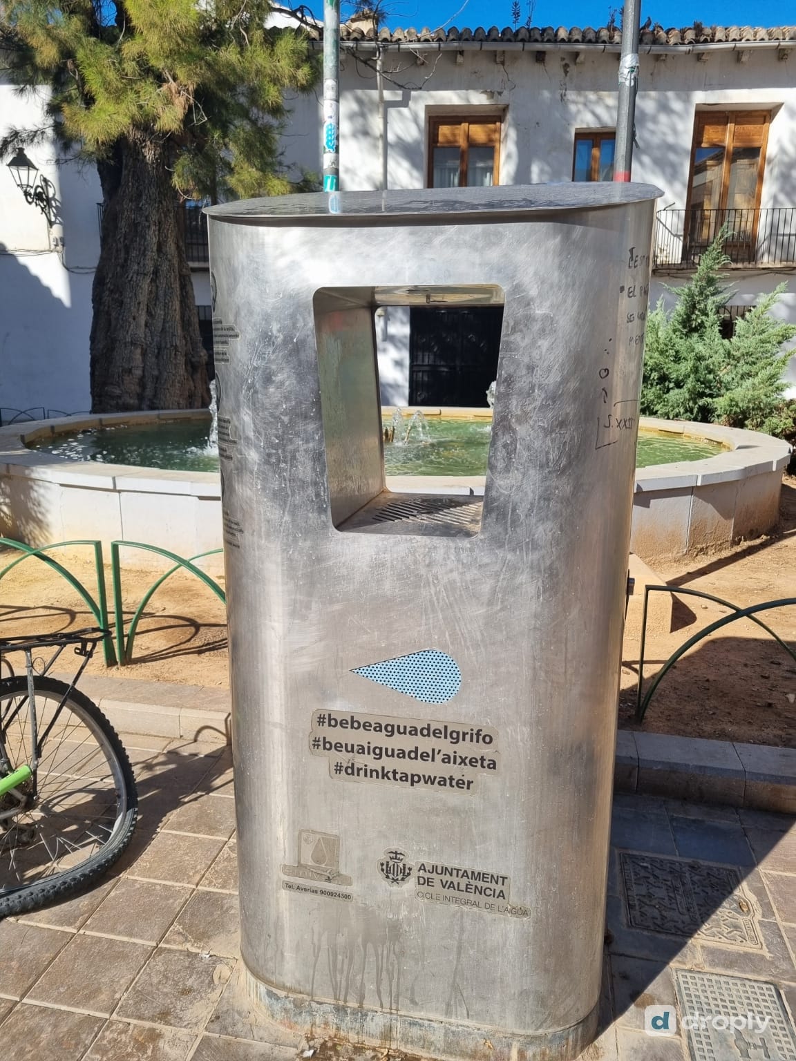 A stainless-steel PUSDAR refill dispenser in Valencia with the hashtags #bebeaguadelgrifo, #beuaiguadelaixeta and #drinktapwater and the logo of the Ajuntament de València, in front of a small square with a fountain