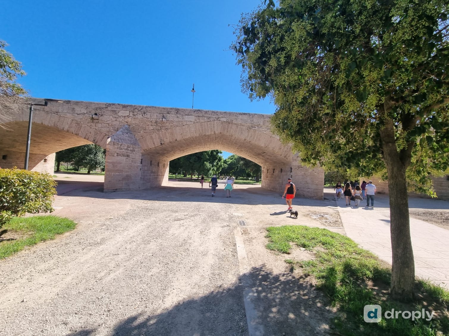 The Turia Garden in Valencia, the former riverbed, with an old stone bridge, trees, and people walking, running and walking a dog on the gravel path