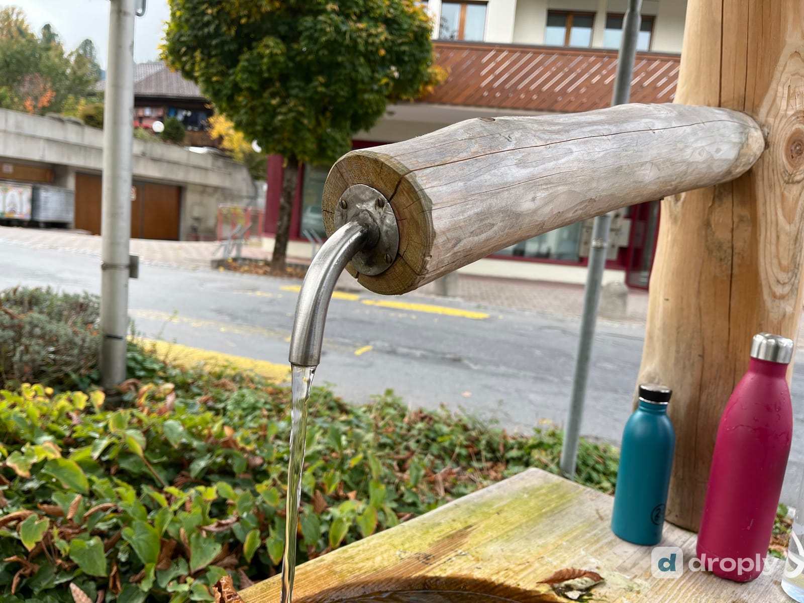 A wooden village fountain running into a trough, with a blue and a pink water bottle standing next to it, in Mannried near Zweisimmen